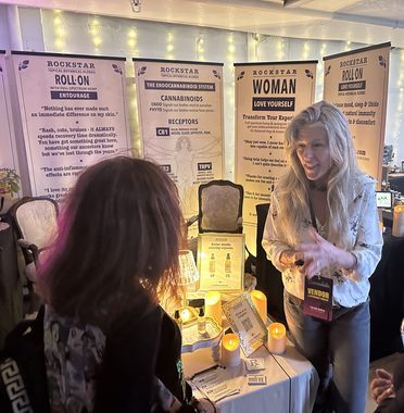 Heather Reilly Hiemstra explains the endocannabinoid system to a visitor at the Rockstar Blends booth, the CB1, CB2 and TRPV receptor diagram and Vitruvian figure on the banner behind her, Cannadelic Night Market.