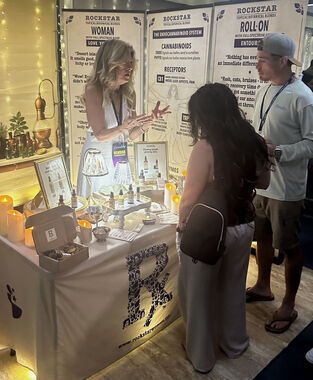 Heather Reilly Hiemstra explains the endocannabinoid system to two visitors at the Rockstar Blends booth, the table draped with the Rockstar "R" logo and displaying Rockstar Woman and Roll-On Entourage products at Cannadelic Night Market.