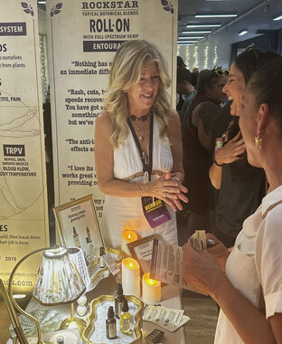 Heather Reilly Hiemstra talks and laughs with two attendees at the Rockstar Blends booth, the candlelit table set with botanical oils, a small lamp, and an Instagram card beneath the Roll-On Entourage banner at Cannadelic Night Market.