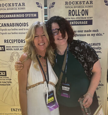 Heather Reilly Hiemstra talks with two attendees at the Rockstar Blends booth, the table set with botanical oil bottles, candles, and product signage beneath the Roll-On Entourage banner at Cannadelic Night Market.